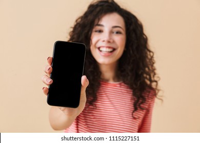 Cheerful young girl with dark curly hair showing blank screen mobile phone isolated over beige background