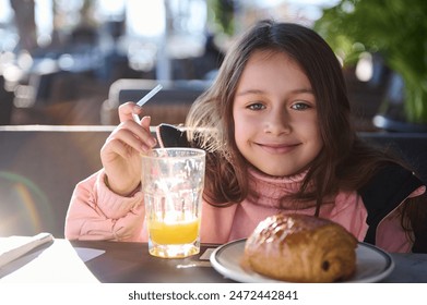 A cheerful young girl enjoys a breakfast of orange juice and a pastry at an outdoor cafe. The peaceful morning atmosphere and her joyful expression create a warm and inviting scene. - Powered by Shutterstock