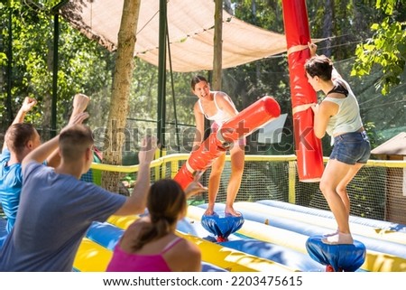 Cheerful young girl with female friend having fun on inflatable gladiator fight arena in outdoor amusement park in summer..