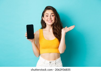 Cheerful young girl introduce your app or store, showing emtpy smartphone screen and smiling at camera, standing against blue background