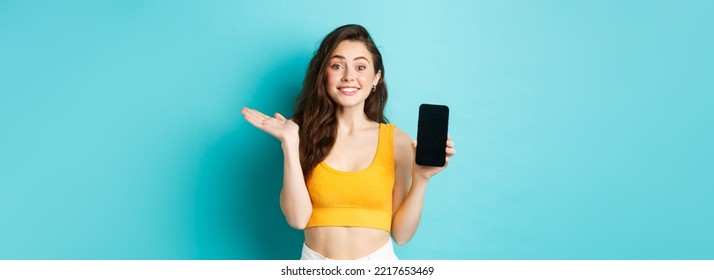 Cheerful young girl introduce your app or store, showing emtpy smartphone screen and smiling at camera, standing against blue background.