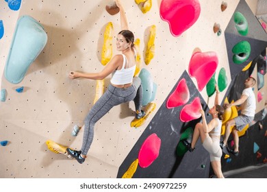 Cheerful young girl practicing bouldering in sports center, climbing colored artificial wall without safety rope, clinging to handholds and footholds, looking at camera with smile.. - Powered by Shutterstock