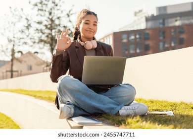 A cheerful young girl sits cross-legged on the grass with her laptop on her lap, wearing headphones around her neck, and giving a friendly wave with her left hand - Powered by Shutterstock