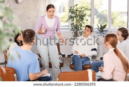 Cheerful young girl student discussing learning assignment with friendly coursemates gathered around table in classroom
