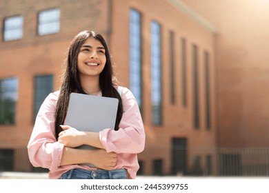 Cheerful young girl student holding a closed laptop, looking upwards thoughtfully beside a university building - Powered by Shutterstock