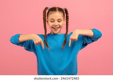 A cheerful young girl with two long braided pigtails is wearing a bright blue sweater. She stands against a solid pink backdrop, playfully pointing down with both her index fingers - Powered by Shutterstock