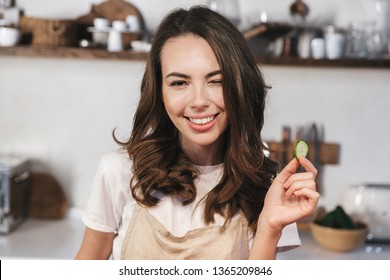 Cheerful young girl wearing apron sitting at the kitchen at home, holding slice of cucumber