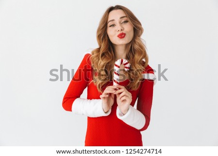 Cheerful young girl wearing Christmas dress standing isolated over white background, eating Christmas candy treats