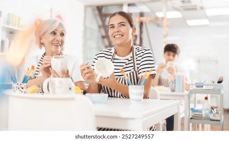 Cheerful young girl working together with elderly mother in family pottery studio, chatting and laughing carefree while creating unique paint designs on ceramic cups.. - Powered by Shutterstock