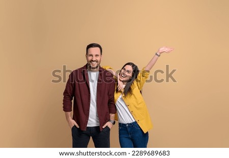 Cheerful young girlfriend with hand raised screaming and leaning on handsome boyfriend. Portrait of attractive couple dressed in shirts posing happily over beige background