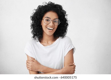 Cheerful Young Good Looking Woman With Clean Dark Skin And Black Shaggy Hair Posing Indoors With Crossed Arms, Smiling Broadly With Her White Straight Teeth, Laughing At Good Joke, Wearing Casual Top