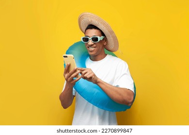 cheerful young guy afro american with inflatable swim ring uses smartphone on yellow isolated background, man in summer on beach is typing on phone, concept of travel and summer holidays