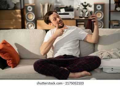 Cheerful Young Guy Communicates On Skype With His Friends. He Is Sitting On The Couch And Holding Phone. On The Sofa Is Plaid And Pizza Boxes. Music Is Playing In The Background.