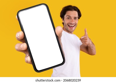 Cheerful young guy holding smartphone with white empty screen, pointing at blank display for mobile application advertisement or your text, gadget mockup template, isolated orange studio background - Shutterstock ID 2068020239