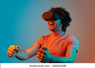 Cheerful Young Guy In VR Glasses Keeping Hands On Invisible Steering Wheel While Driving Race Car In Video Game Under Blue And Orange Illumination