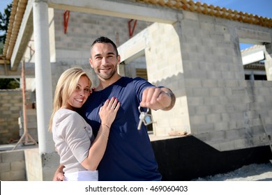 Cheerful Young Happy Couple Holding Home Keys In Front Of Construction Site Of New House