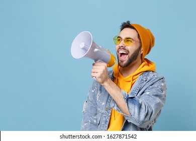 Cheerful young hipster guy in fashion jeans denim clothes posing isolated on pastel blue background studio portrait. People lifestyle concept. Mock up copy space. Looking aside, scream in megaphone