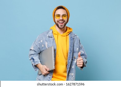 Cheerful young hipster guy in fashion jeans denim clothes posing isolated on pastel blue background in studio. People lifestyle concept. Mock up copy space. Hold laptop pc computer, showing thumb up