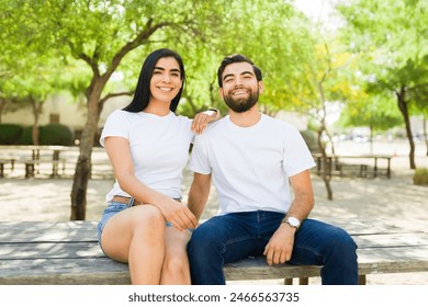 Cheerful young hispanic couple wearing white t-shirt mockups enjoy a sunny day together in a park, showing bright smiles - Powered by Shutterstock