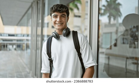 Cheerful young hispanic male student standing outdoors enjoying campus life, confidently wearing headphones and backpack, radiating positive energy and joy at the university. - Powered by Shutterstock