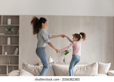 Cheerful Young Hispanic Woman Having Fun Dancing To Disco Music Jumping On Cozy Couch With Adorable Child Daughter, Enjoying Carefree Leisure Weekend Time Activity Together In Modern Living Room.