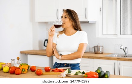 Cheerful young Hispanic woman relaxing after work at home, enjoying glass of red wine while preparing vegetable salad for dinner - Powered by Shutterstock