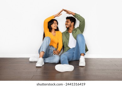 Cheerful young Indian couple in casual clothing sitting on the floor, smiling, and creating roof shape with their raised hands against white wall - Powered by Shutterstock