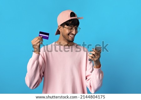 Cheerful young indian guy hipster holding modern cell phone and credit card, shopping or banking online, using mobile application, blue studio background, copy space. E-commerce concept