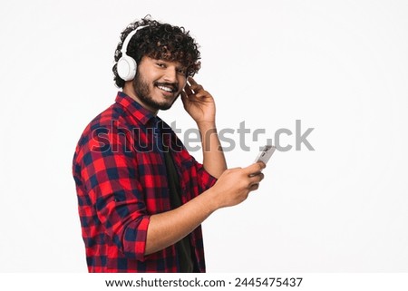 Cheerful young Indian man in casual clothes listening to the music in headphones isolated over white background. Hindi fan enjoying podcast e-book playlist song