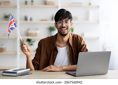 Cheerful Young Indian Man English Teacher Showing Flag Of UK, Sitting In Front Of Laptop At Office, Teaching Via Online Courses, Leading Language School, Copy Space. Online Education Concept