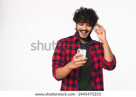Cheerful young Indian man listening to the music in headphones isolated over white background. Hindi boy choosing music audio track on cellphone in mobile application