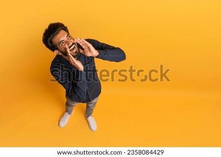 Cheerful Young Indian Man Making Announcement, Keeping Hands Near Mouth, Above Shot Of Funny Eastern Guy Sharing News Or Information While Standing Isolated On Yellow Background, Copy Space