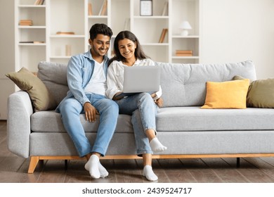 Cheerful young indian spouses comfortably sitting on modern gray couch, looking at laptop screen together with bright, engaging smiles in cozy room, copy space - Powered by Shutterstock