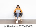 Cheerful young indian woman using laptop computer and wireless headphones, sitting in armchair over white blank wall background, watching movie podcast, have video call, copy space