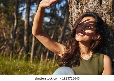 Cheerful Young Lady Blocking The Sun With Her Hand To Shield Her Face From Its Rays All While Leaning Against A Tree