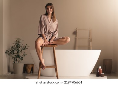 Cheerful Young Lady In Elegant Silk Dressing Gown Sitting On Wooden Stool In Stylish Luxury Bathroom Against Modern Ceramic Bathtub, Ready To Take Bath, Having Beauty And Spa Procedures