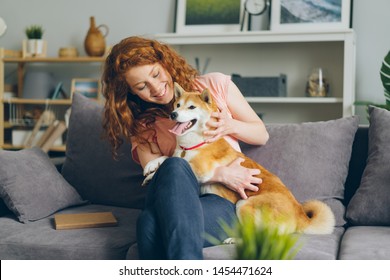 Cheerful young lady happy dog owner is stroking beautiful shiba inu puppy on couch in flat sitting together smiling. Lifestyle, love and friendship concept.