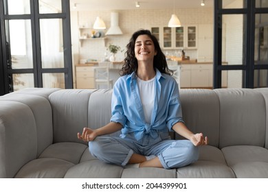 Cheerful young lady sitting on comfy couch, meditating with closed eyes at home, full length. Peaceful millennial woman doing yoga in lotus pose on sofa, finding inner balance, managing stress indoors
