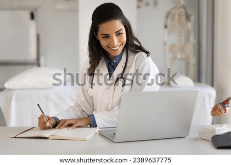 Cheerful young Latin doctor woman talking to patient on video call, taking notes, writing complaints, smiling, laughing. Happy medical practitioner learning on Internet, watching course