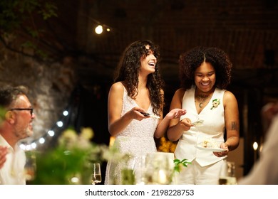 Cheerful young lesbian brides in white wedding attire spreading pieces of cut big cake among guests sitting by served festive table - Shutterstock ID 2198228753