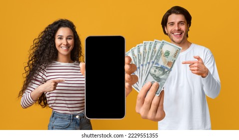 Cheerful young lovers man and woman gambling on Internet, using cell phone with black empty screen, pointing at smartphone, bunch of dollars, yellow studio background, mockup. Online bet concept - Shutterstock ID 2229551355