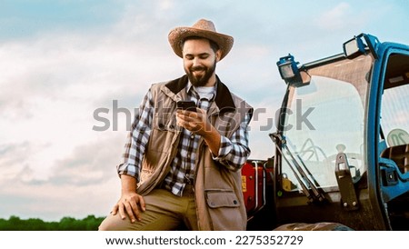 Cheerful young male driver at farm standing at tractor, tapping and texting on smartphone and resting. Farming concept. Happy man farmer having rest and using mobile phone for chatting. Outside.