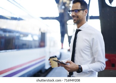 Cheerful Young Male Entrepreneur In Formal Clothes Standing On Platform Waiting For Train Getting To Job In Morning, Smiling Businessman Holding Portable Pc And Coffee To Go On Railway Station