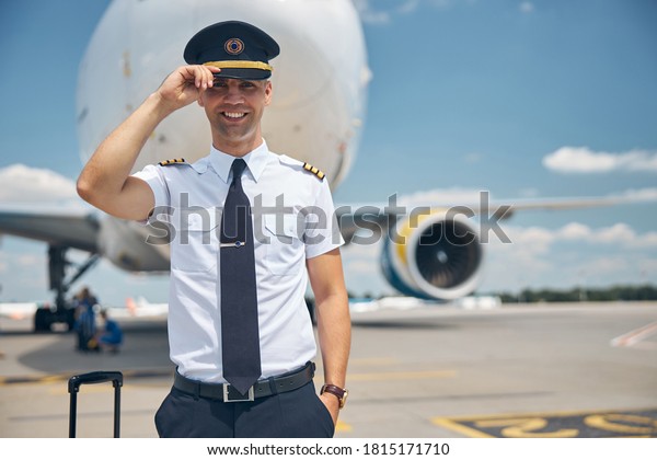 Cheerful\
young man airline worker touching captain hat and smiling while\
standing in airfield with airplane on\
background