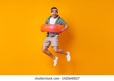 Cheerful Young Man In Cap, Sneakers, Shorts And Shirt Is Having Fun And Jumping With Inflatable Circle On Orange Background