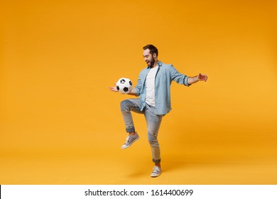 Cheerful Young Man In Casual Blue Shirt Posing Isolated On Yellow Orange Wall Background, Studio Portrait. People Emotions Lifestyle Concept. Mock Up Copy Space. Juggling Bouncing Soccer Ball On Knee