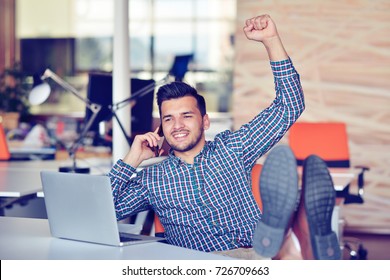 Cheerful young man in casual wear keeping arms raised and looking happy while sitting at the desk in office - Powered by Shutterstock