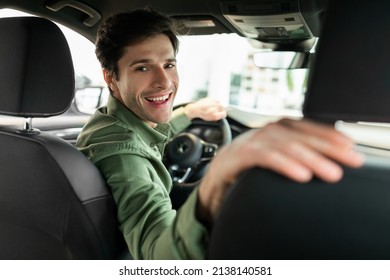Cheerful Young Man Driving New Automobile After Purchase, Sitting In Driver's Seat Of Car At Dealership. Happy Millennial Guy Looking Back And Smiling, Buying Modern Auto At Showroom