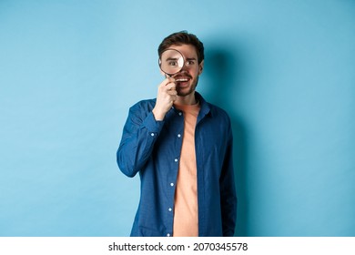Cheerful young man found something good, smiling and looking through magnifying glass, standing on blue background
