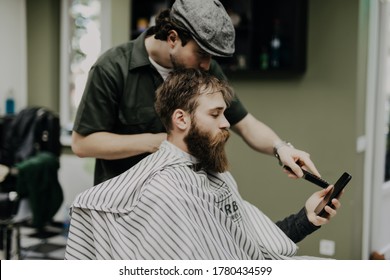 Cheerful Young Man Getting Haircut By Hairdresser While Sitting In Chair. Holding Phone.